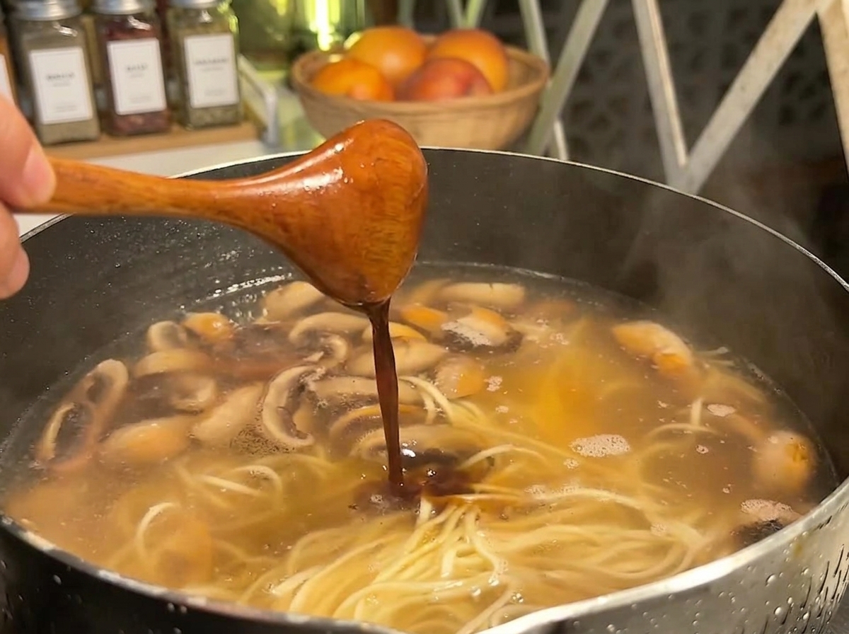Salsa scura versata da un cucchiaio di legno in una pentola di noodles, brodo e funghi affettati.