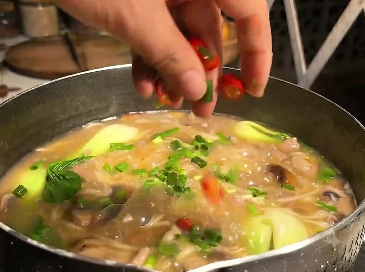 Una mano che cosparge anelli di peperoncino rosso affettato su una pentola bollente di zuppa di noodles piena di verdure e carne.