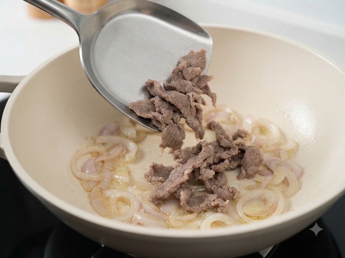 Fette di manzo sbollentate trasferite con una spatola in una padella bianca con cipolle affettate saltate.