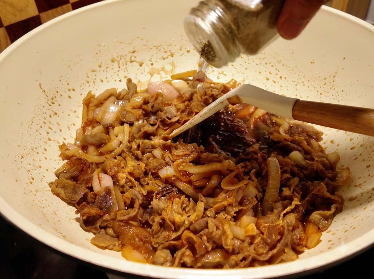 Pepe nero macinato da un contenitore di vetro su manzo e cipolle che sobbollono nella salsa in padella.