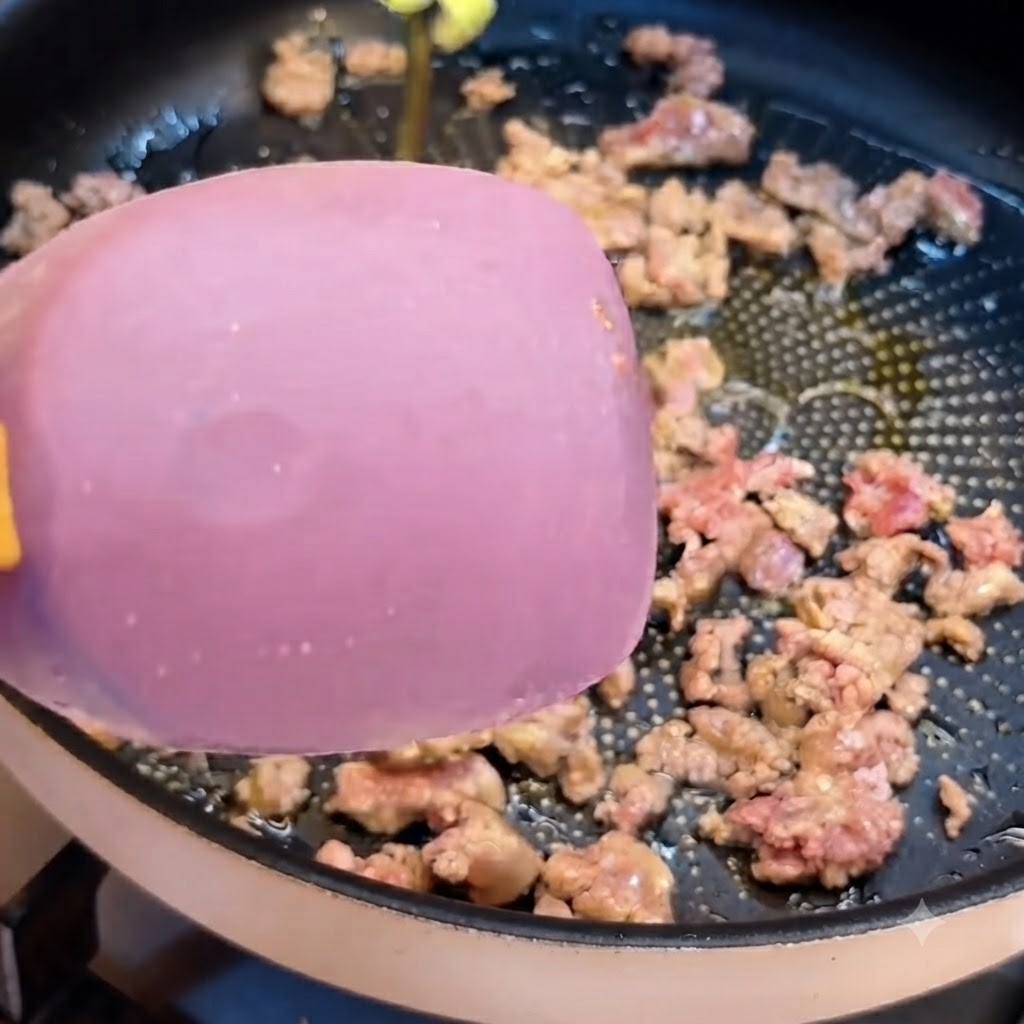 Una spatola rosa che si muove attraverso la carne macinata saltata in una padella scura.