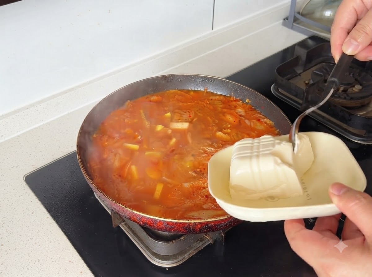 Blocchi di tofu bianco calati in una pentola di zuppa di kimchi rossa.