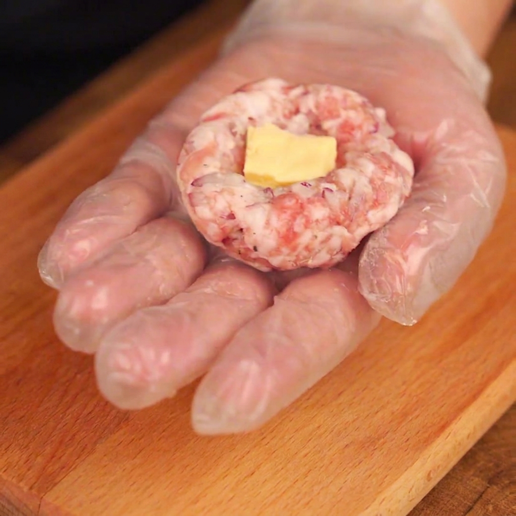 Un pezzetto di formaggio giallo posizionato nel centro cavo di una polpetta di maiale modellata in una mano guantata.