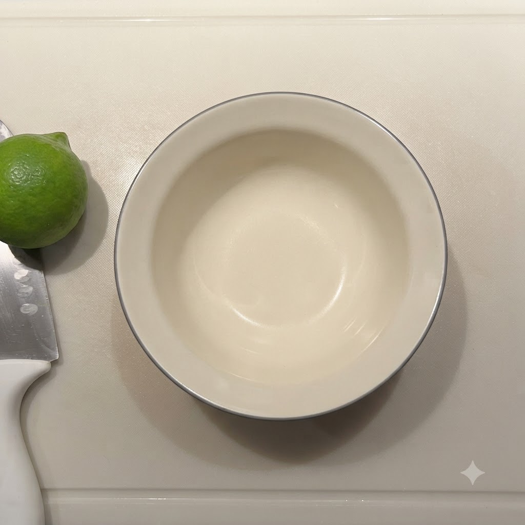 Una piccola ciotola di ceramica vuota accanto a un lime fresco e un coltello su un tagliere.