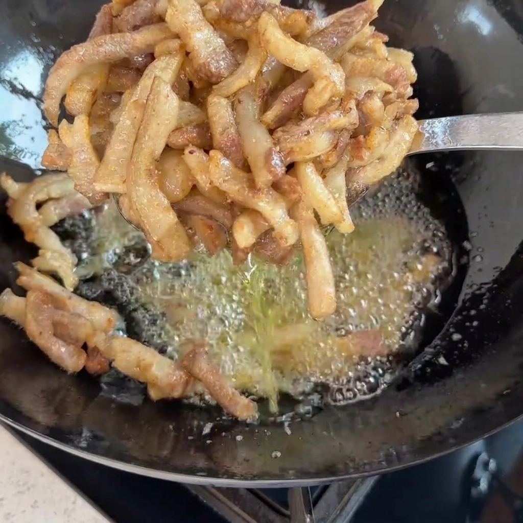 Un mestolo forato in metallo che solleva gli straccetti di maiale appena fritti e dorati su un wok di olio bollente.