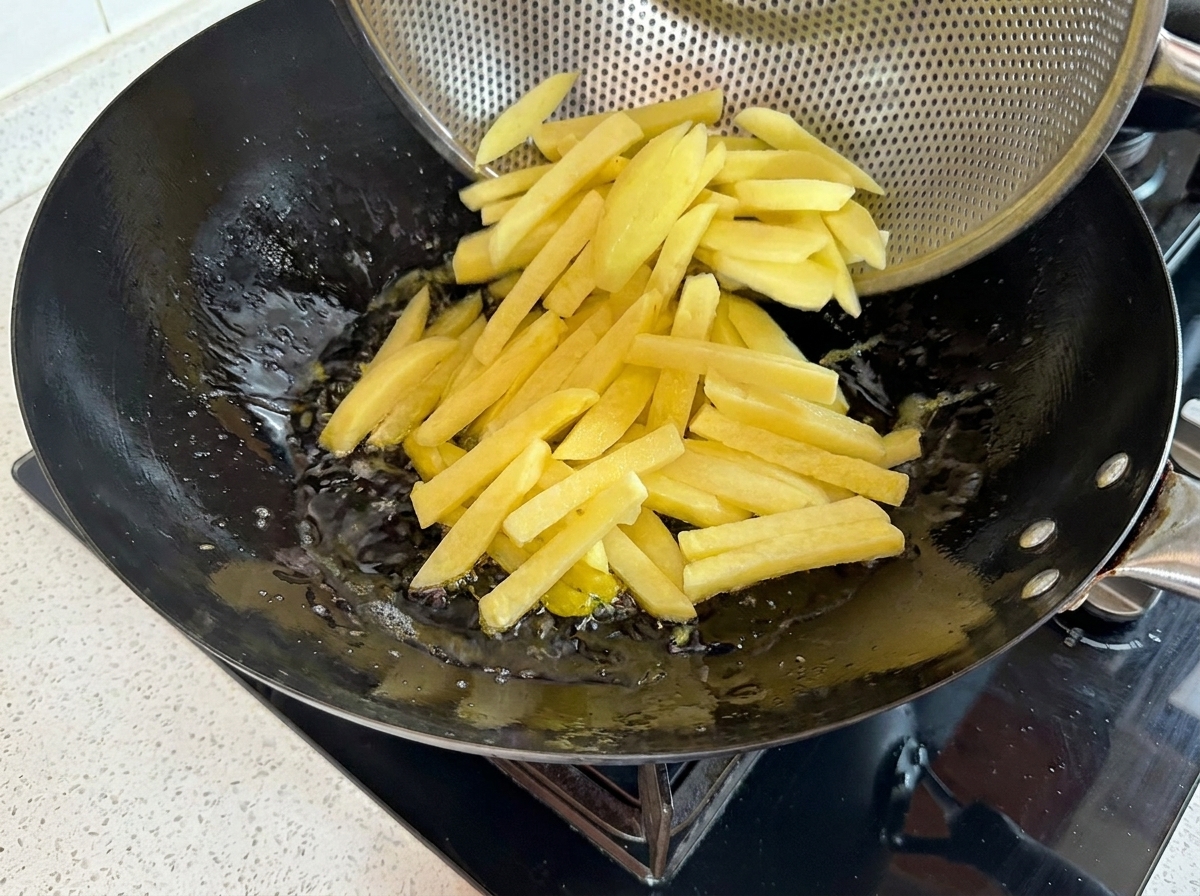 Bastoncini di patate fresche che vengono versati da un colino in metallo in un wok di olio bollente.