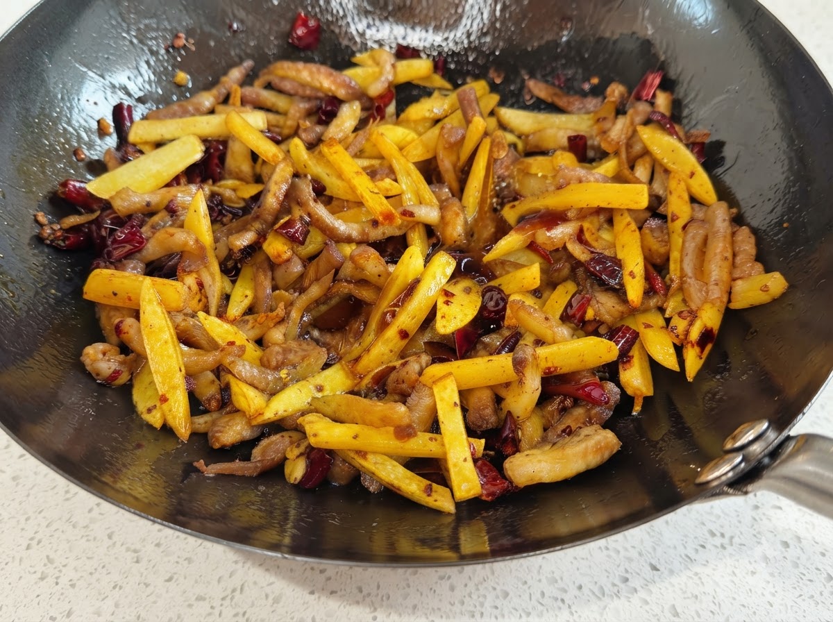 Un wok pieno di straccetti di patate e maiale che brillano di salsa di soia e sono spolverati di peperoncino rosso e spezie marroni.