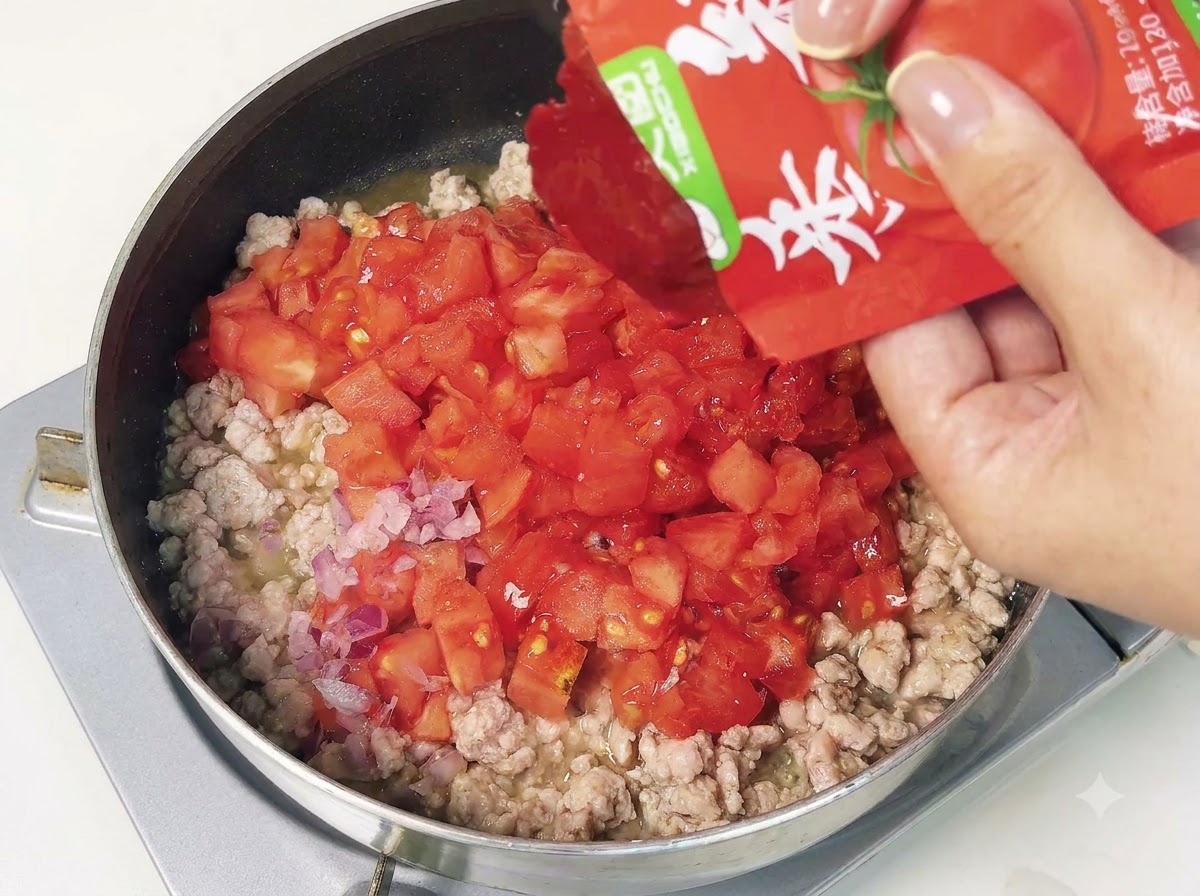 Un pacchetto di concentrato di pomodoro svuotato in una padella con carne macinata e pomodori a cubetti.