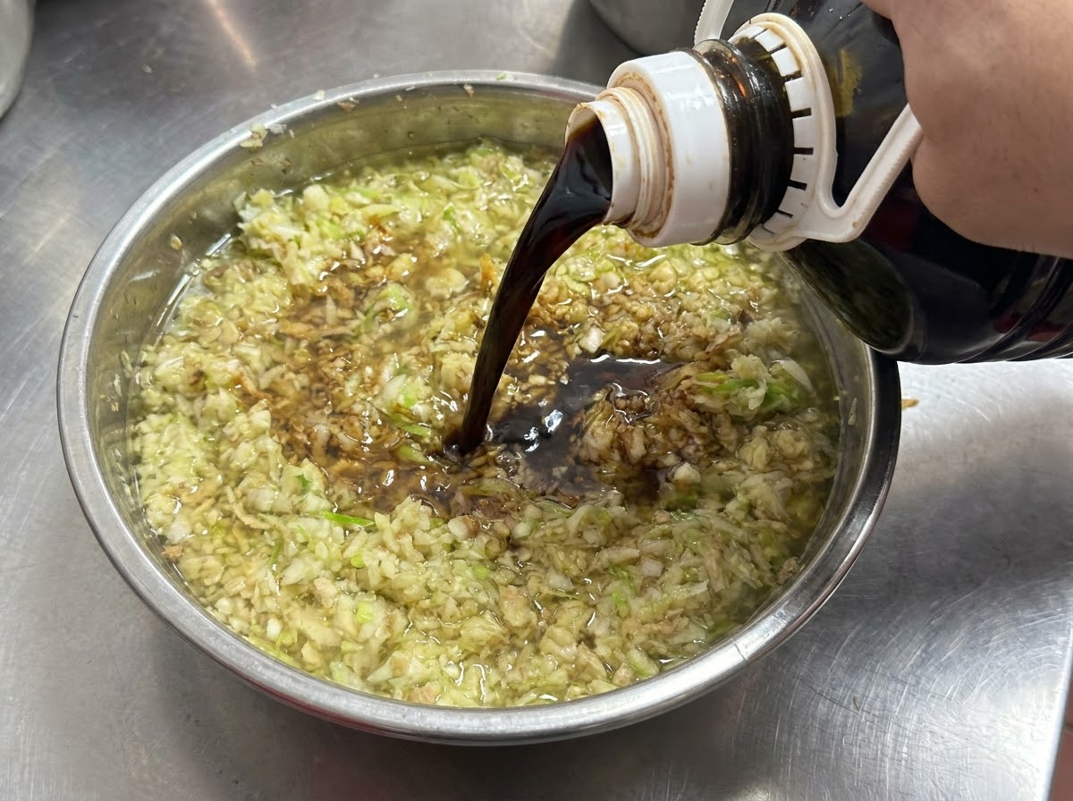 Salsa di soia scura versata da una bottiglia in una ciotola piena di aglio tritato e cipollotti.