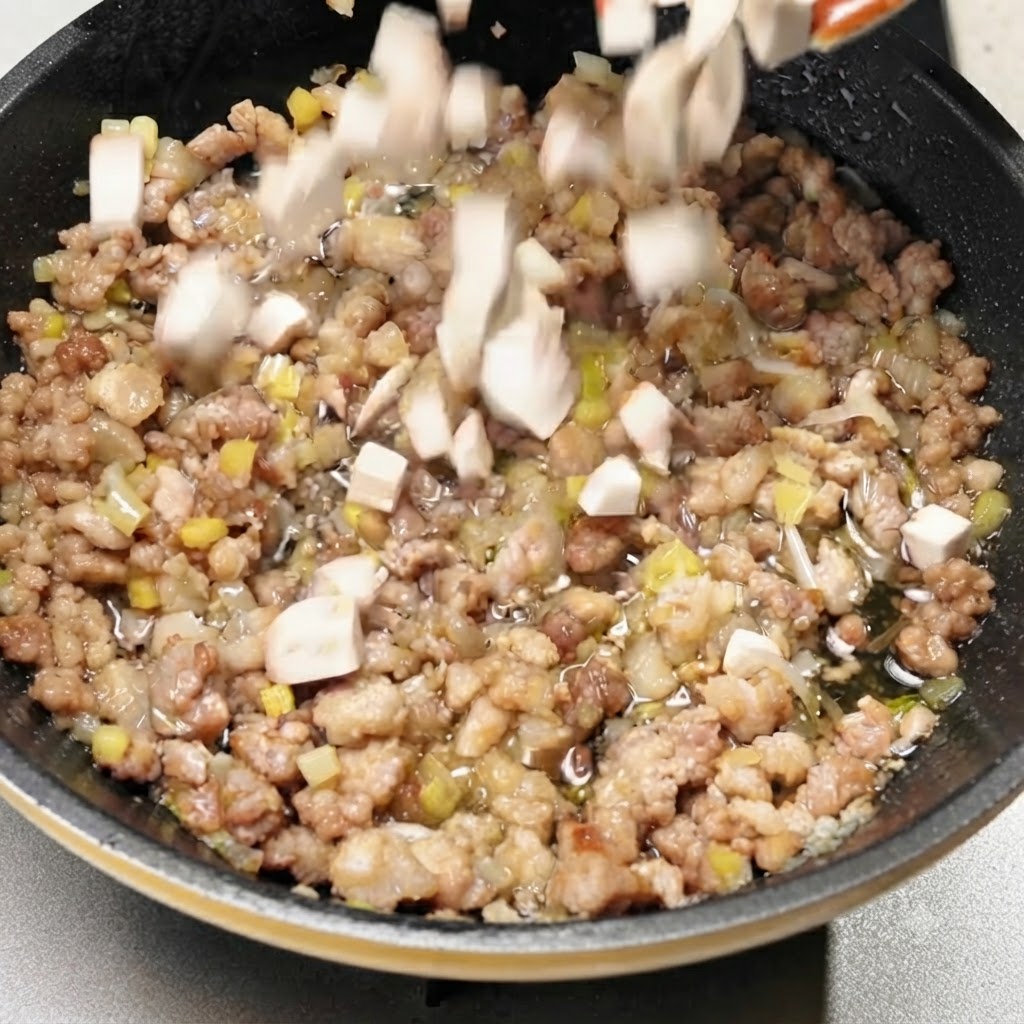 Cubetti di funghi tagliati a dadini che cadono nella padella piena di maiale tritato cotto e aromi.