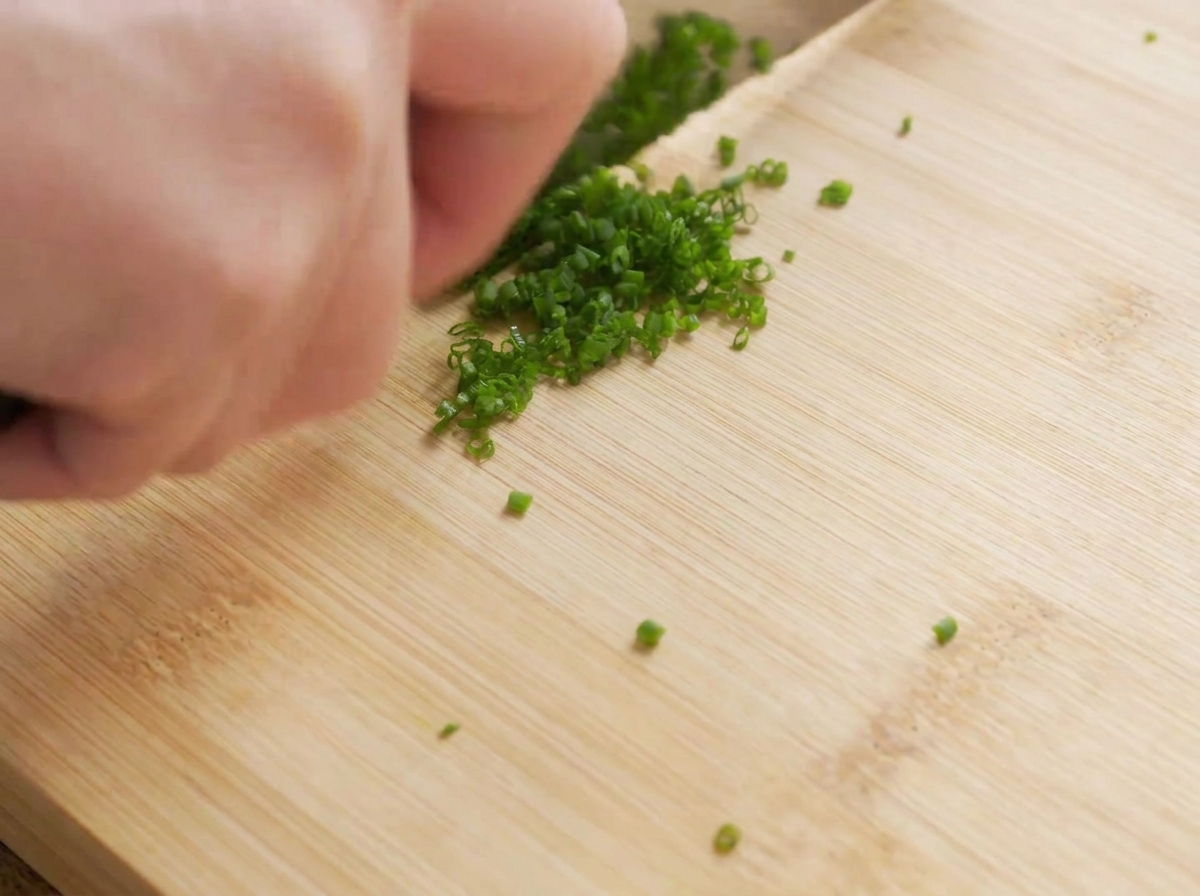 Un primo piano di un coltello che trita finemente lerba cipollina fresca su un tagliere di legno.