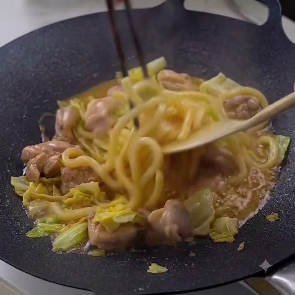 Bacchette e una spatola di legno che mescolano noodles udon fumanti, pollo e cavolo in una padella scura.