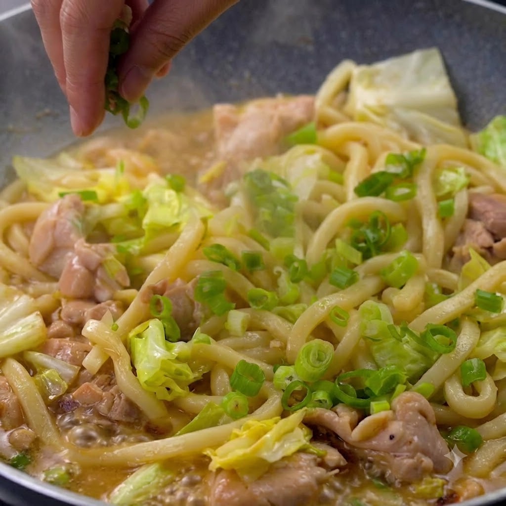 Una mano che cosparge cipollotti tritati vivaci su una padella di noodles udon e pezzi di pollo fumanti.